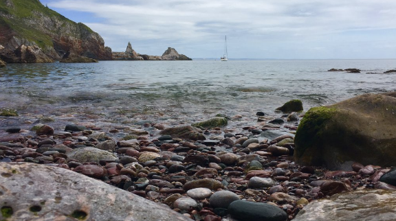 a pebbled beach looking out onto the sea which has a sailing boat by some rocks on it