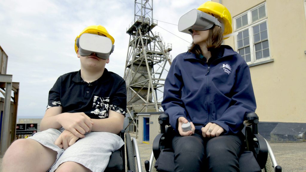 A man and a woman in wheelchairs with VR headsets on their face. There is a large mining structure in the background.