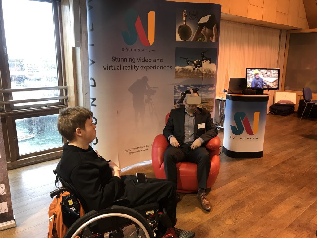 a man in a chair with a VR headset on ands another in a wheelchair watching