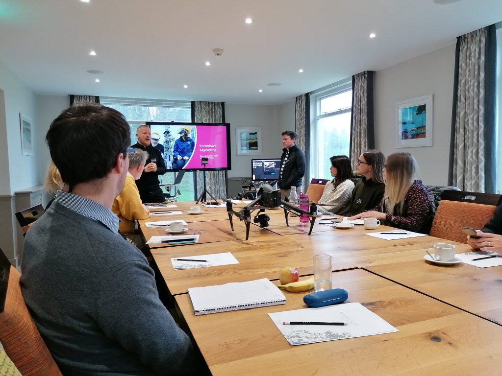 people sat around a table with a drone on it watching two men present a powerpoint slide