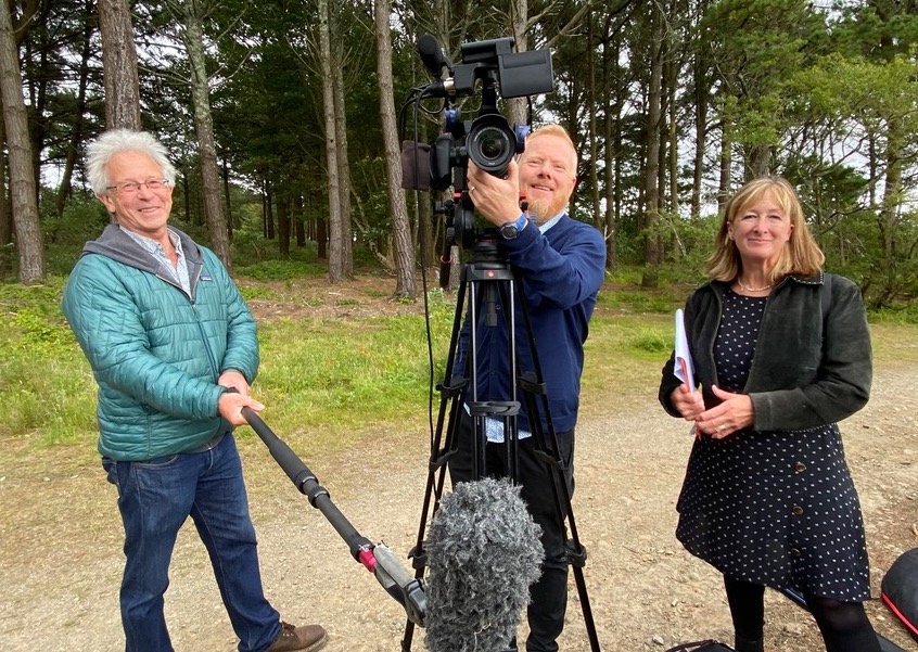 Three people stood in a forest smiling at the camera, a man holding a large microphone, another man a video production camera and a female holding some papers.