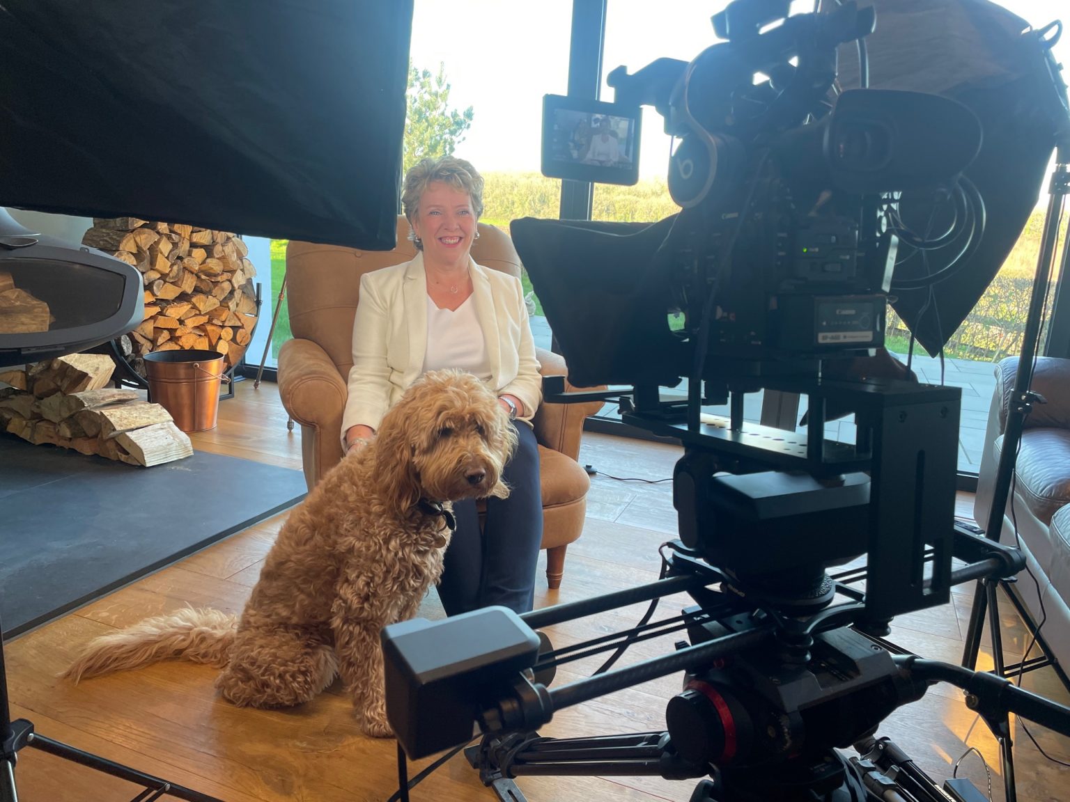 A Woman smiling towards a camera who is accompanied by a brown dog.