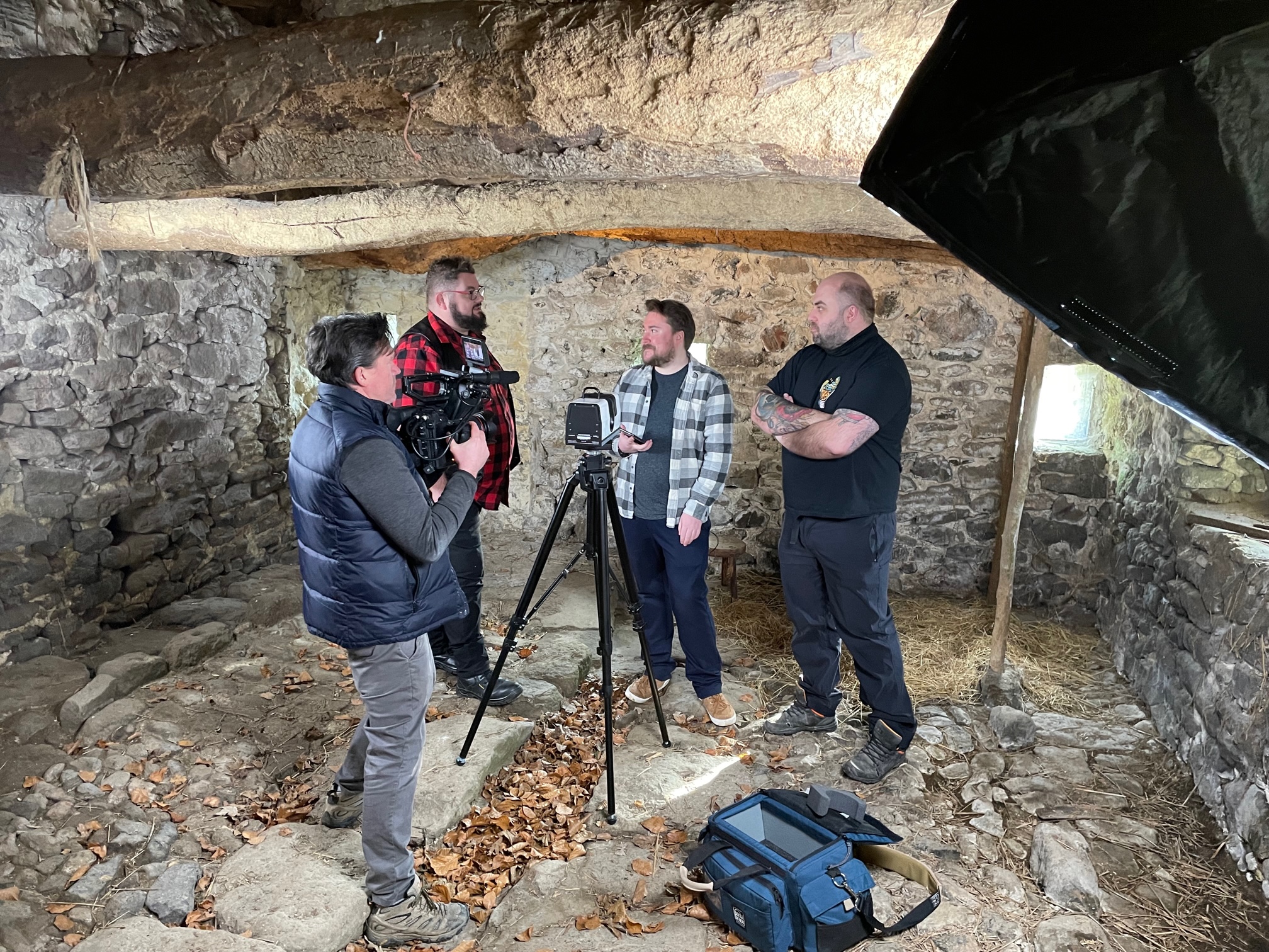 A man holding a camera filming three other men. They are in a cobblestone building.