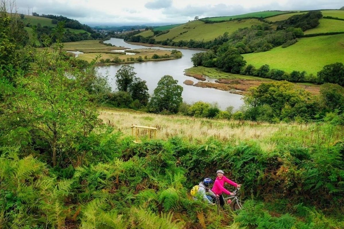 a man and woman cycling towards the left hand corner of the image. The are surrounded by trees hedges and a river with two valleys either side