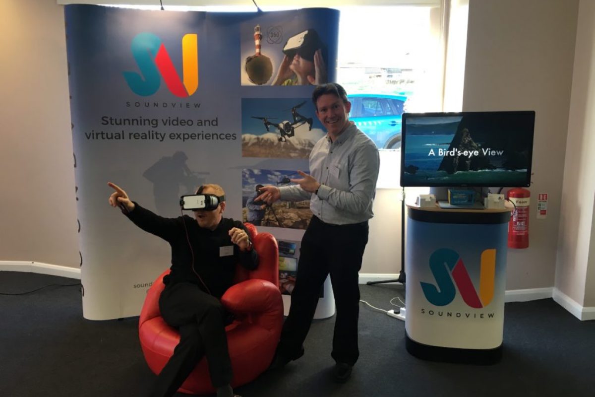 two men posing funnily in front of a business show setup wearing VR headsets
