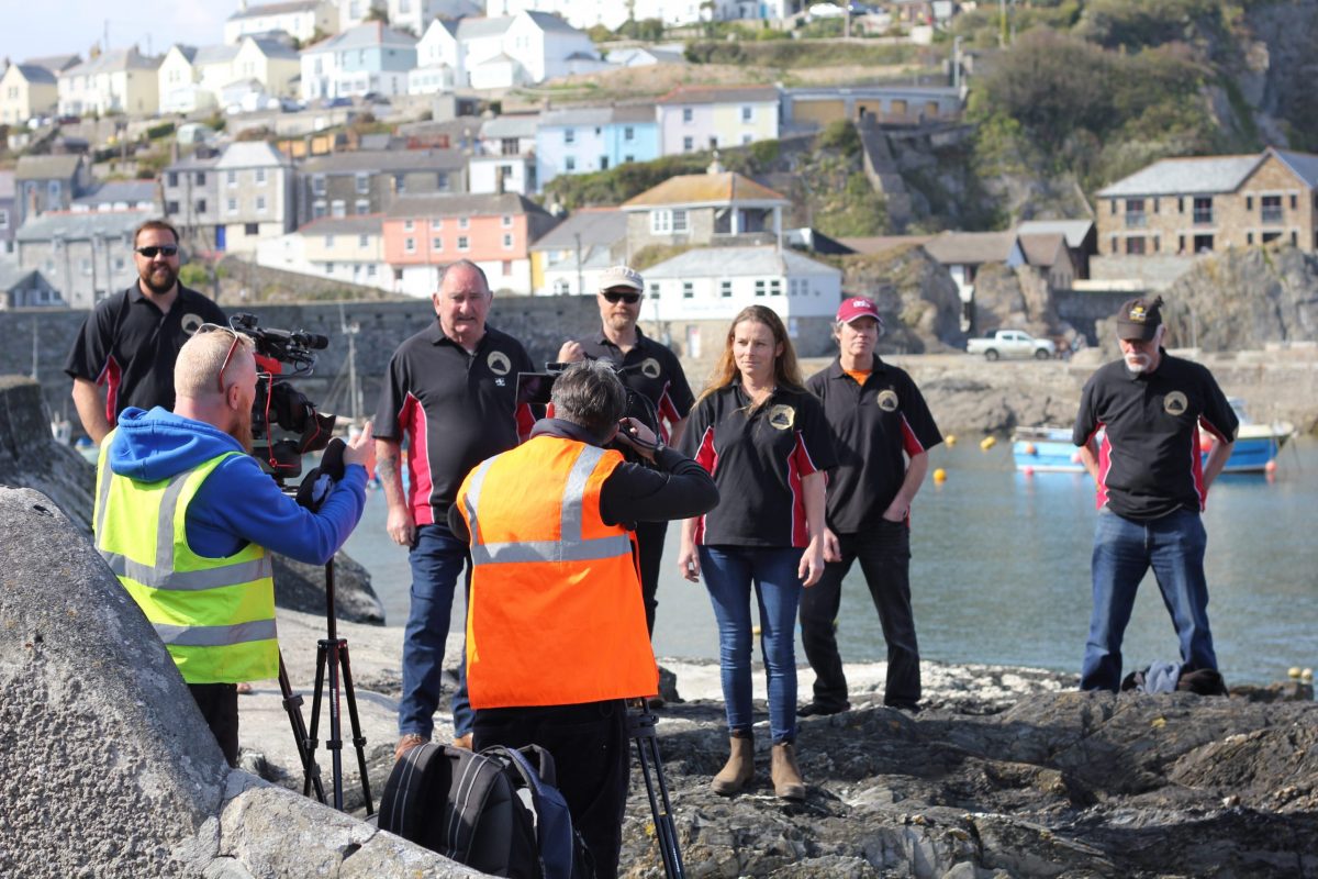 A group of people stood in front of a harbour being filmed by two men in high vis and video production equipment.