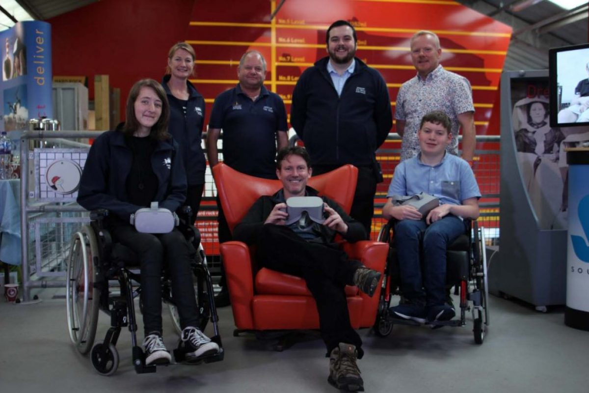 seven people posing for a picture smiling. There are two wheelchair users and one sat on a large red chair with four people behind them.