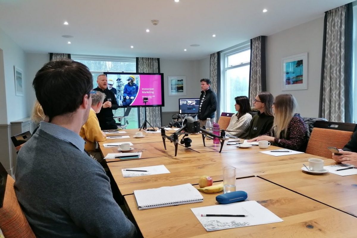 people sat around a table with a drone on it watching two men present a powerpoint slide
